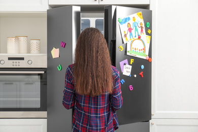 Woman opening refrigerator door with child's drawings, notes and magnets in kitchen