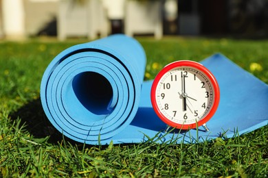 Photo of Alarm clock and fitness mat on green grass outdoors. Morning exercise