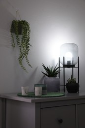 Stylish lamp, candles and green plants on grey chest of drawers indoors. Interior design