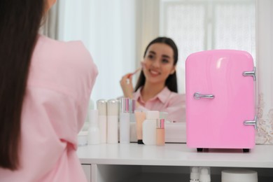 Woman doing face massage at dressing table with cosmetic refrigerator indoors