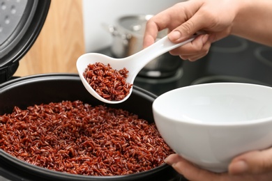 Woman putting brown rice into bowl from multi cooker in kitchen, closeup