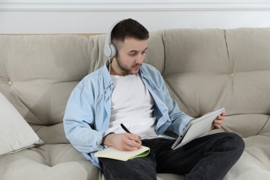 Photo of Young man with headphones using modern tablet for studying on sofa at home. Distance learning