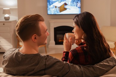 Photo of Happy lovely couple with hot drinks spending time together near fireplace at home, back view