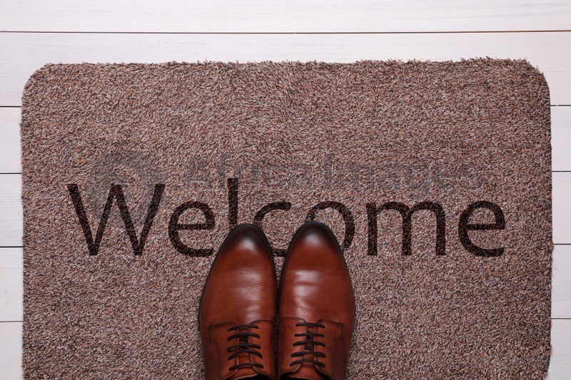 Door mat with word WELCOME and shoes on white wooden floor, top view