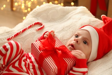 Cute little baby in Santa hat holding Christmas present on knitted blanket