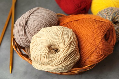 Different balls of woolen knitting yarns in wicker basket and needles on grey background, closeup