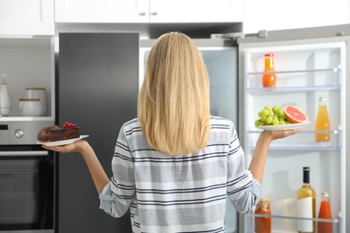 Choice concept. Woman holding plates with fruits and sweets near refrigerator in kitchen, back view