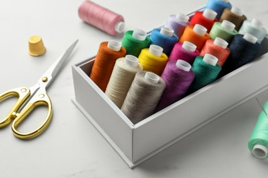 Box with colorful sewing threads, scissors and thimble on white marble table