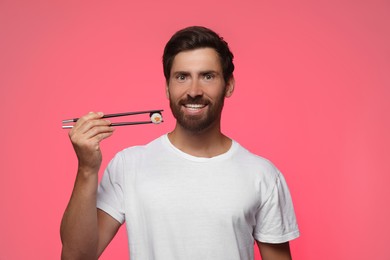 Photo of Happy man holding tasty sushi roll with chopsticks on pink background