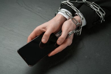 Man holding smartphone in chained hands at black table, closeup. Internet addiction
