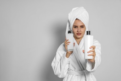 Photo of Beautiful young woman in bathrobe holding bottles of shampoo on light grey background. Space for text