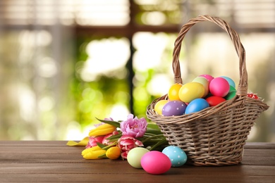 Colorful Easter eggs in wicker basket and tulips on wooden table indoors, space for text