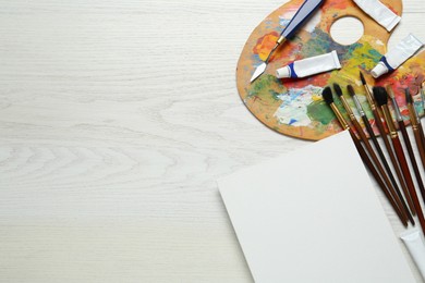 Blank canvas, tools, palette and paints on white wooden table, flat lay. Space for text