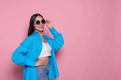 Attractive happy woman touching fashionable sunglasses against pink background. Space for text