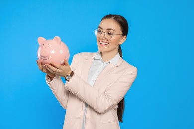 Happy young businesswoman with piggy bank on light blue background
