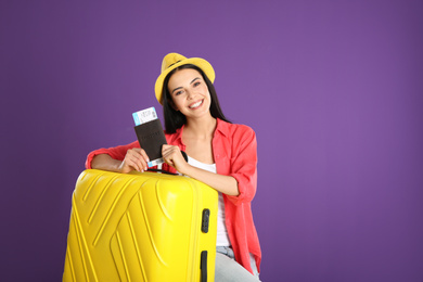 Beautiful woman with suitcase and ticket in passport for summer trip on purple background. Vacation travel