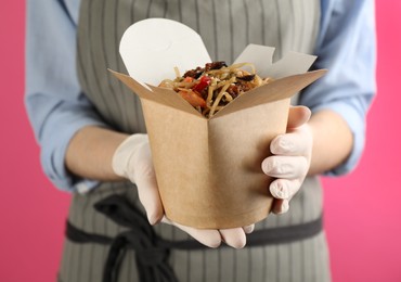 Chef holding box of wok noodles with seafood on pink background, closeup