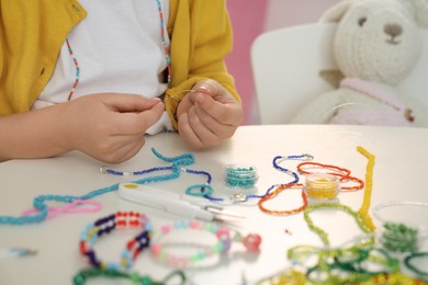Little girl making beaded jewelry at table in room, closeup
