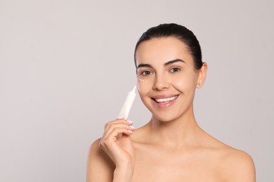 Beautiful young woman applying gel on skin under eye against light grey background, space for text