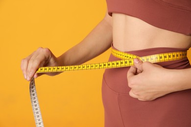 Woman in sportswear measuring waist with tape on yellow background, closeup