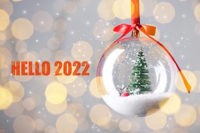 Text Hello 2022 and beautiful transparent Christmas ornament against blurred festive lights, bokeh effect