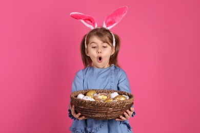 Surprised little girl with bunny ears holding wicker basket full of Easter eggs on pink background