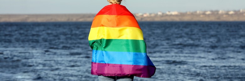 Woman wrapped in bright LGBT flag near river, back view. Banner design