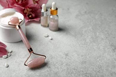 Photo of Natural rose quartz face roller, cosmetic products and flowers on grey background. Space for text