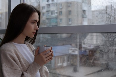 Melancholic young woman with drink looking out of window on rainy day, space for text. Loneliness concept