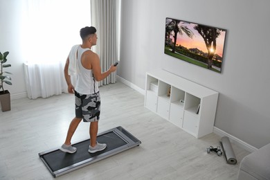 Sporty man with remote control training on walking treadmill and watching TV at home