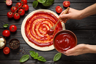 Woman spreading tomato sauce onto pizza crust on dark wooden table, top view
