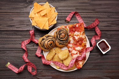 Photo of Plate of different unhealthy food and measuring tape on wooden table, flat lay. Weight loss concept