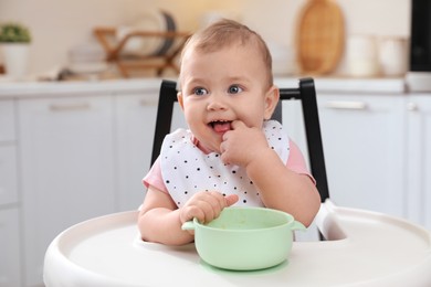 Cute little baby with bowl in high chair at kitchen