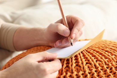 Man writing message in greeting card on orange
knitted pouf, closeup