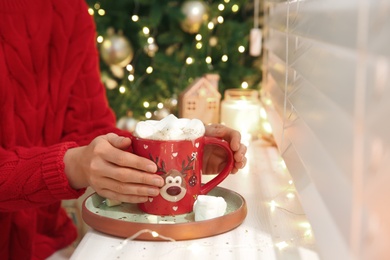 Woman with delicious marshmallow cocoa near window indoors, closeup. Christmas celebration