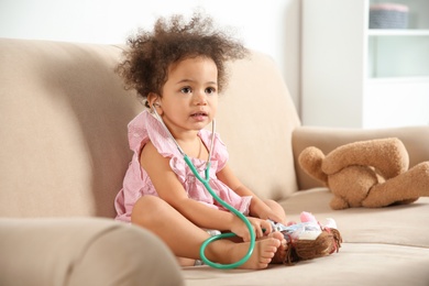 Cute African American child imagining herself as doctor while playing with stethoscope and doll on couch at home