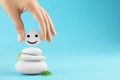 Woman putting stone with drawn happy face on stack against light blue background, closeup and space for text. Zen concept