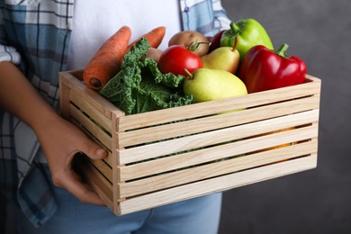 Photo of Farmer holding wooden crate filled with fresh vegetables and fruits on grey background, closeup