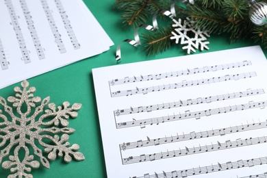 Christmas music sheets near fir tree branches and decorative snowflakes on green background, closeup