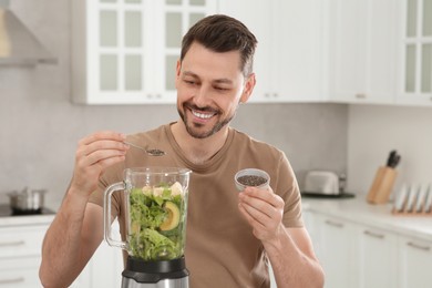 Happy man adding chia seeds into blender with ingredients for smoothie in kitchen