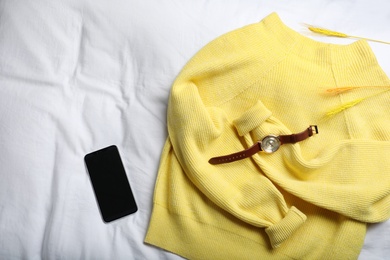 Yellow warm sweater with smartphone and wristwatch on bedsheet, flat lay