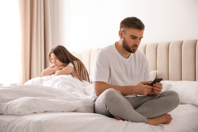 Photo of Young man preferring smartphone over his girlfriend on bed at home. Relationship problems