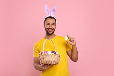 Photo of Happy African American man in bunny ears headband holding wicker basket with Easter eggs on pink background