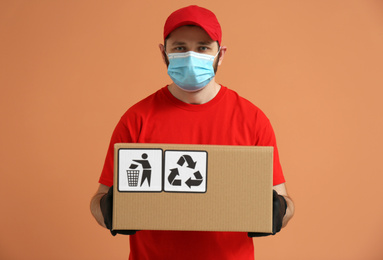 Courier in mask holding cardboard box with different packaging symbols on orange background. Parcel delivery