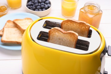 Yellow toaster with roasted bread, glasses of juice, blueberries and jam on white wooden table, closeup