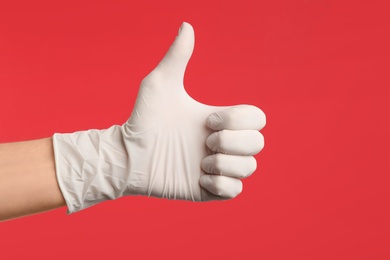 Person in medical gloves showing thumb up on red background, closeup of hand