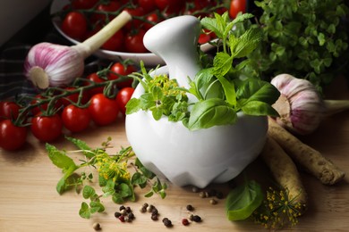 Photo of Mortar with fresh herbs near garlic, horseradish roots, black peppercorns and cherry tomatoes on wooden table, closeup