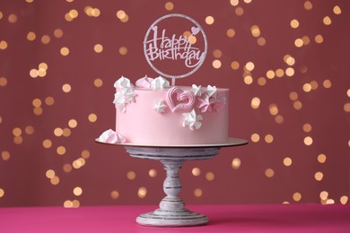 Beautifully decorated birthday cake on pink table against blurred festive lights