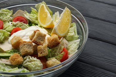 Bowl of delicious salad with Chinese cabbage, lemon, tomatoes and bread croutons on black wooden table, closeup