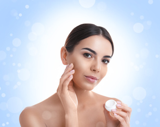 Young woman applying facial cosmetic cream on light blue background with bokeh effect